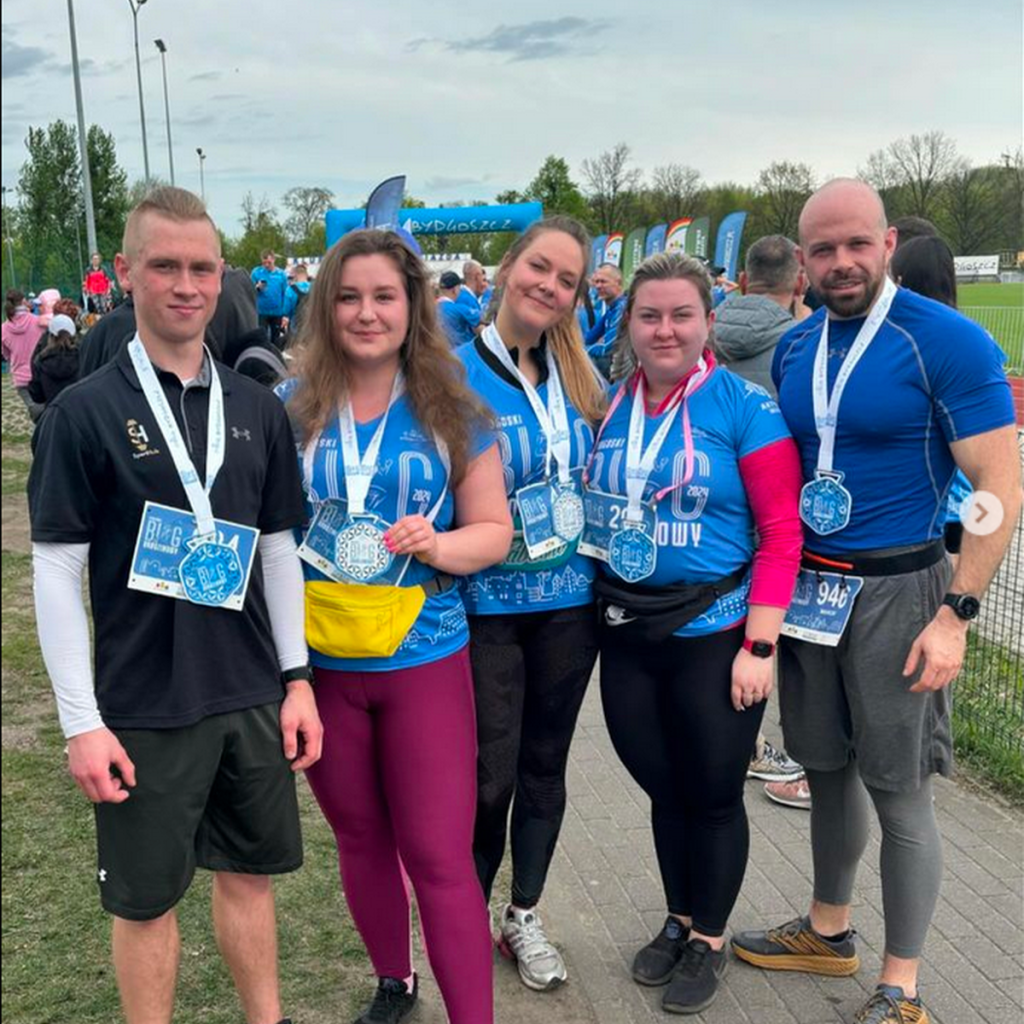 Grupa pięciu osób pozuje do zdjęcia po ukończonym biegu, trzymając na szyjach medale pamiątkowe. Ubrani w koszulki sportowe, głównie w odcieniach niebieskiego z logo wydarzenia, wyglądają na zadowolonych i dumni ze swojego osiągnięcia. W tle widoczny jest teren biegowy z flagami i uczestnikami wydarzenia.